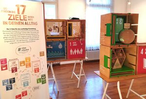 Ausstellungstafel mit dem Text 17 Ziele in deinem Alltag. daneben zwei Tische mit sechs Holzkisten. Vorne offen. 