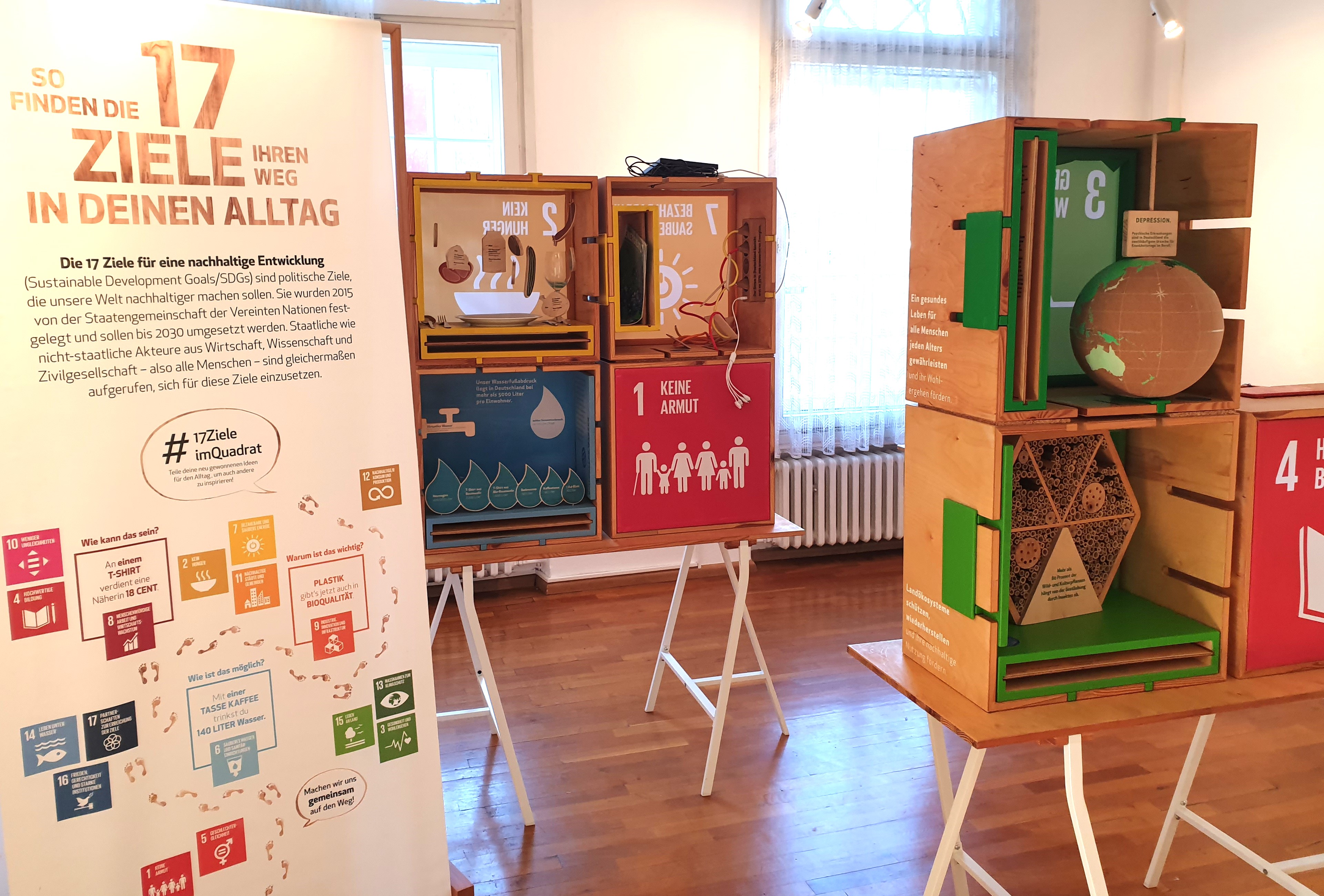 Ausstellungstafel mit dem Text 17 Ziele in deinem Alltag. daneben zwei Tische mit sechs Holzkisten. Vorne offen. Ausstellungstafel mit dem Text 17 Ziele in deinem Alltag. daneben zwei Tische mit sechs Holzkisten. Vorne offen.